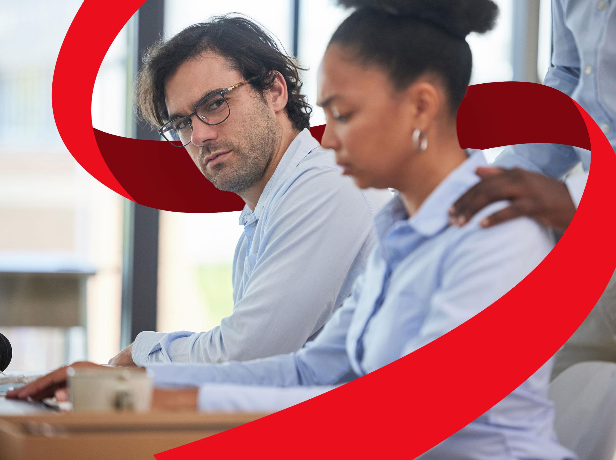 Business professionals seated at a desk.  One is touching another's shoulder and they appear uncomfortable.