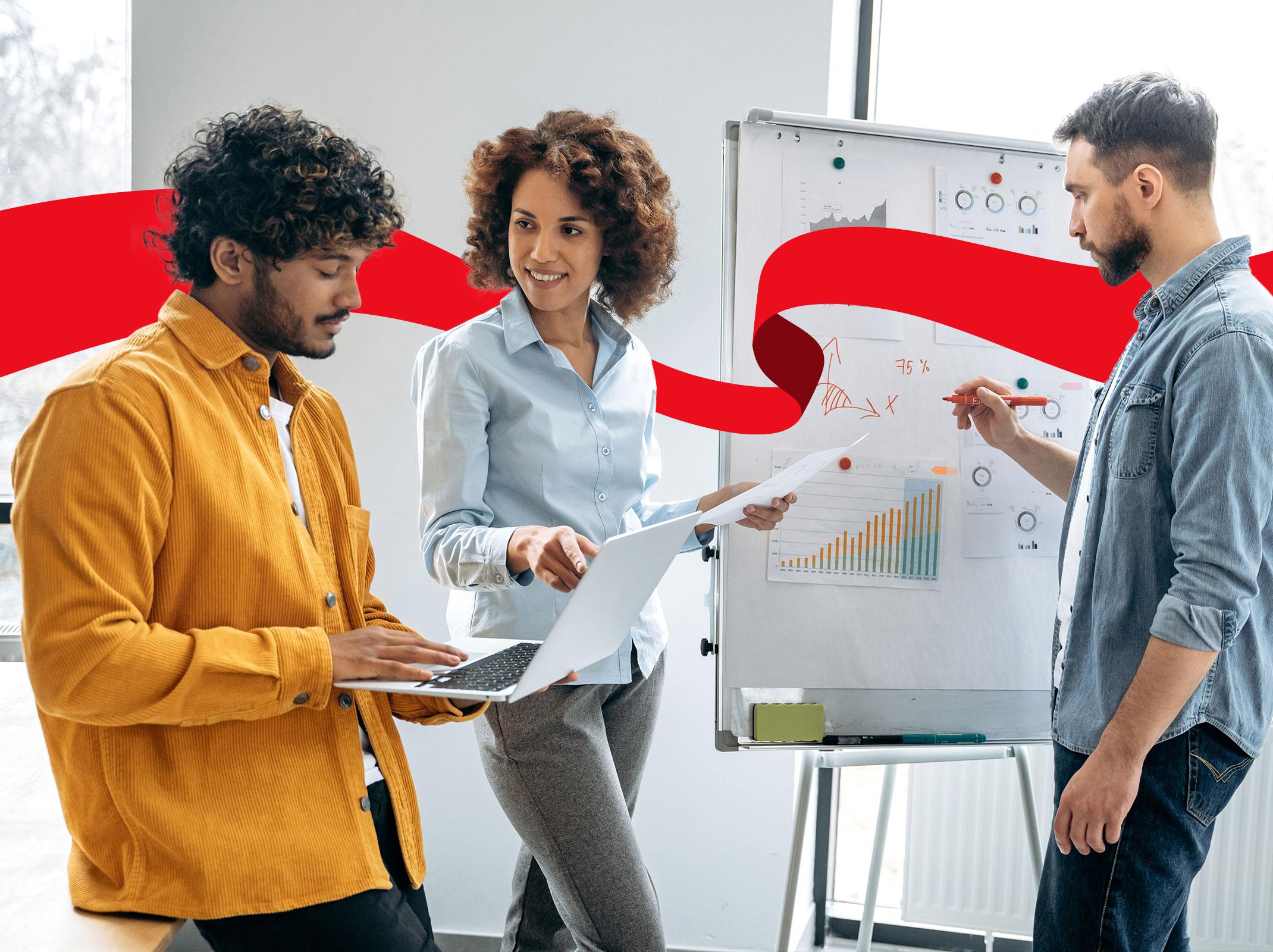 Three marketing professionals reviewing information on a whiteboard and on a laptop.  They are in an office setting.