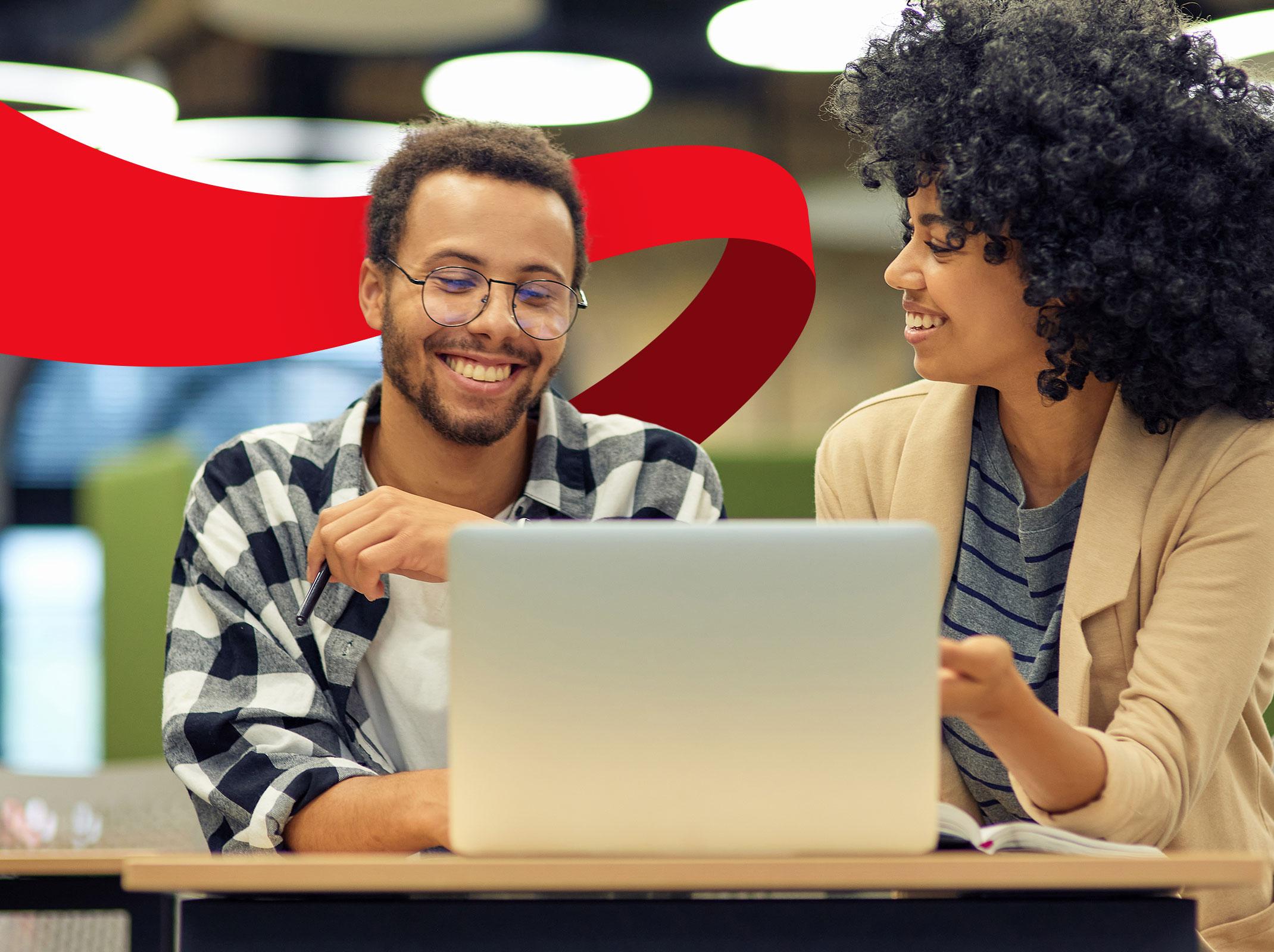 Two business professionals sitting in front of a laptop.  One is pointing at the laptop and both are smiling in a business setting.