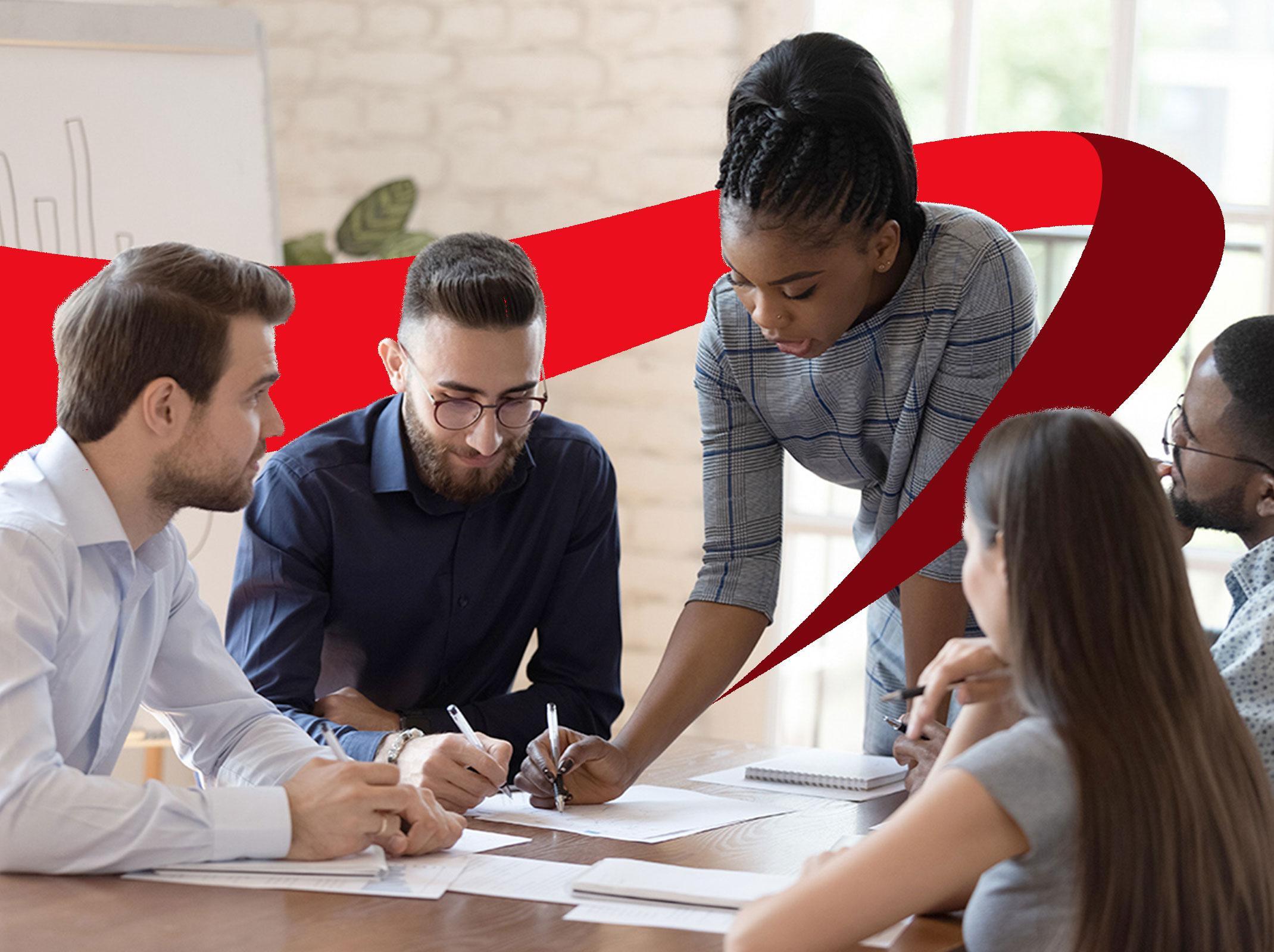 Four employees sitting at conference room table with manager standing up leaning over table writing on a piece of paper.