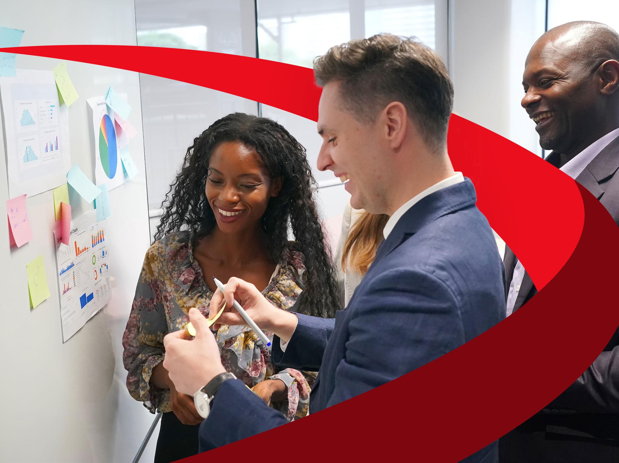 Three business professionals working around a whiteboard.  They are talking and adding notes to the board.  They are in an office setting.
