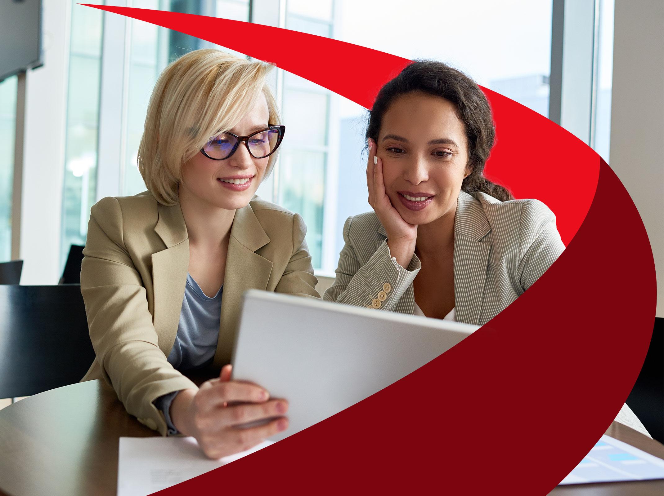 Two female leaders reviewing information on a tablet. They are in a business setting and are sitting at a desk.